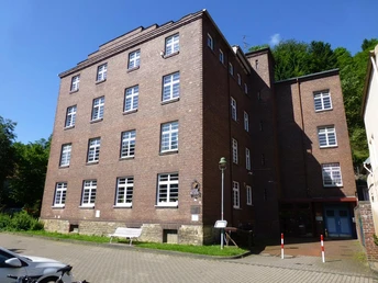 Backsteingebäude mit mehreren Stockwerken, rechteckige Fenster, gepflasterter Vorplatz, grüner Hintergrund.