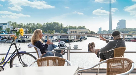 Zwei Personen entspannen auf einer Terrasse mit Blick auf einen Hafen voller Boote, sonniger Tag.