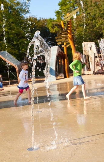 Wasserspielplatz im Maximilianpark Hamm Kinder rennen durch sprudelnde Wasserfontänen eines Spielplatzes, umgeben von Holzspielgeräten.