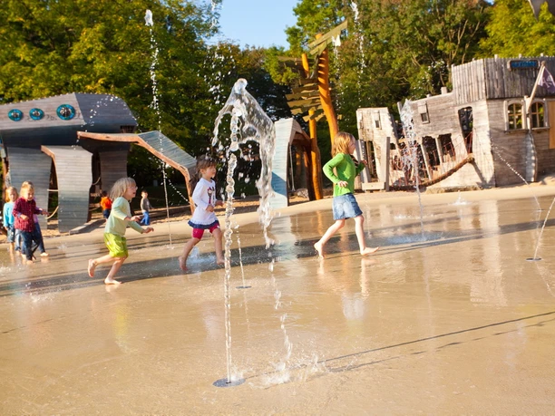 Wasserspielplatz im Maximilianpark Hamm Kinder rennen durch sprudelnde Wasserfontänen eines Spielplatzes, umgeben von Holzspielgeräten.
