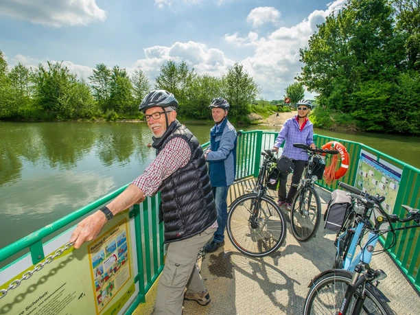 Lippefähre Lupia Personen mit Fahrrädern auf der Lippefähre Lupia überqueren den ruhigen Fluss; umgeben von grünen Bäumen.