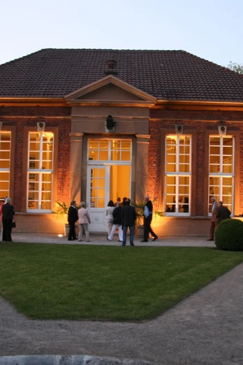 Rotes Backsteingebäude mit großer Fensterfront, abends beleuchtet, Besucher im Vordergrund.