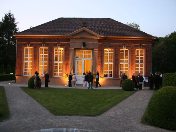 Orangerie Rotes Backsteingebäude mit großer Fensterfront, abends beleuchtet, Besucher im Vordergrund.