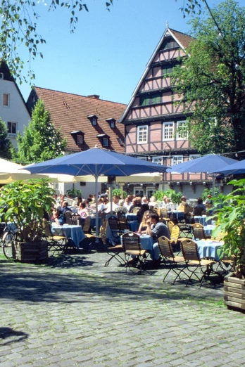 Ein sonniger Tag auf einem Platz, auf dem viele Menschen an Tischen im Freien sitzen und speisen.