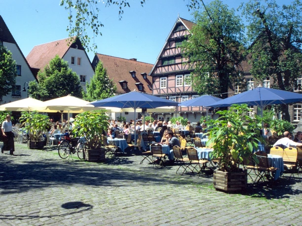 Marktplatz Ein sonniger Tag auf einem Platz, auf dem viele Menschen an Tischen im Freien sitzen und speisen.