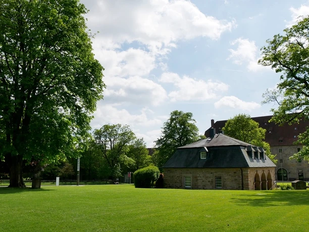 Ein kleines Steingebäude mit Schieferdach steht auf einer grünen Wiese, umgeben von Bäumen.