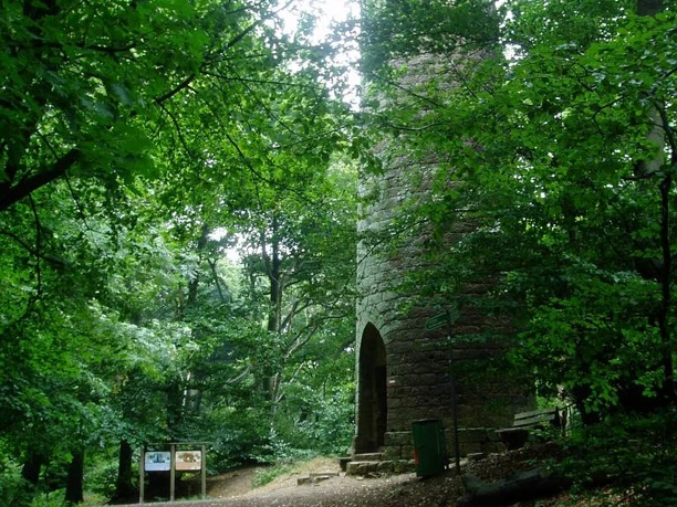 Steinerner Turm im Wald, umgeben von dichtem Blattwerk, mit Informationstafeln am Wegesrand.