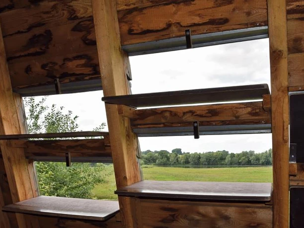 Variable Öffnungen für Vogelbeobachtungen Holzkonstruktion mit mehreren rechteckigen Öffnungen zur Vogelbeobachtung, umgeben von grüner Landschaft.
