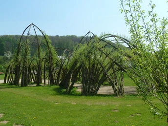 Weidenpalais aus geflochtenen Weidenruten, mit grünen Blättern, auf einer Wiese mit Wald im Hintergrund.