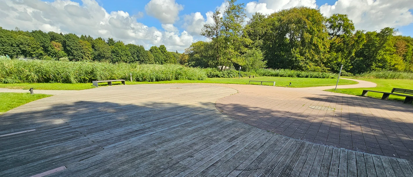 Breite Holz- und Pflasterfläche im Gesundheitspark Speckenbüttel mit Bänken und viel Grün.