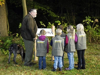 Fünf Kinder in Westen stehen um einen Infotafel, begleitet von einem Erwachsenen und einem Hund im Wald.