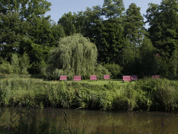 Rote Parkbänke am Wasser vor einer Trauerweide auf der Dalkeinsel in Gütersloh.