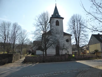 Weißer Kirchenbau, umgeben von einer historischen Mauer mit Baumbestand.