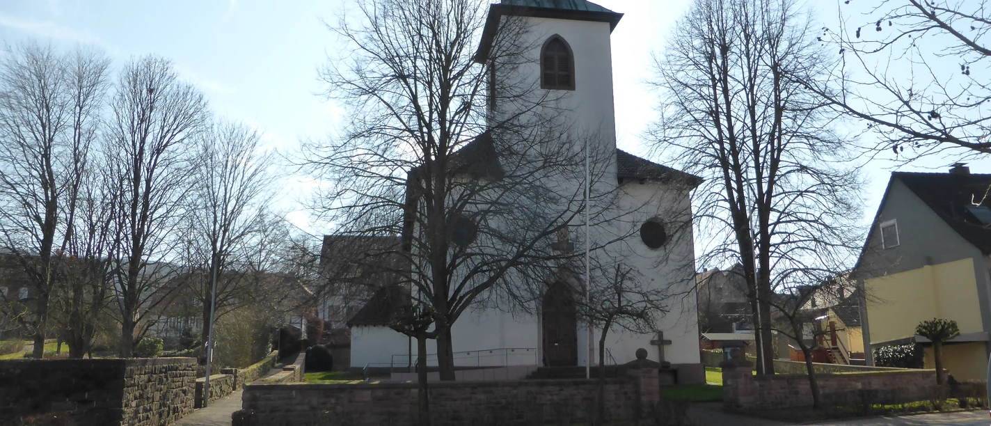 Weißer Kirchenbau, umgeben von einer historischen Mauer mit Baumbestand.