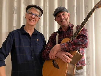 Never give up Eine Frau und ein Mann stehen gemeinsam vor einem Fenster. Der Mann hält eine Akustikgitarre in den Händen.