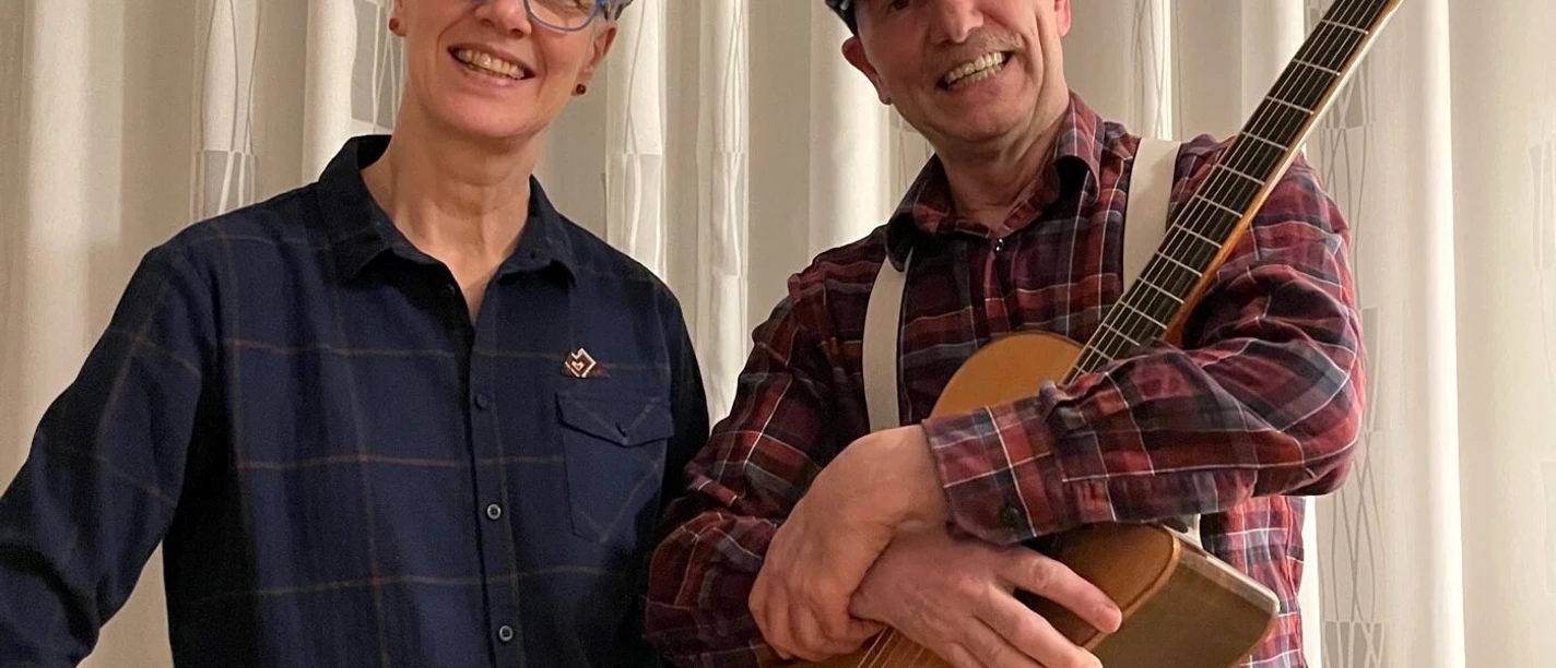 Never give up Eine Frau und ein Mann stehen gemeinsam vor einem Fenster. Der Mann hält eine Akustikgitarre in den Händen.