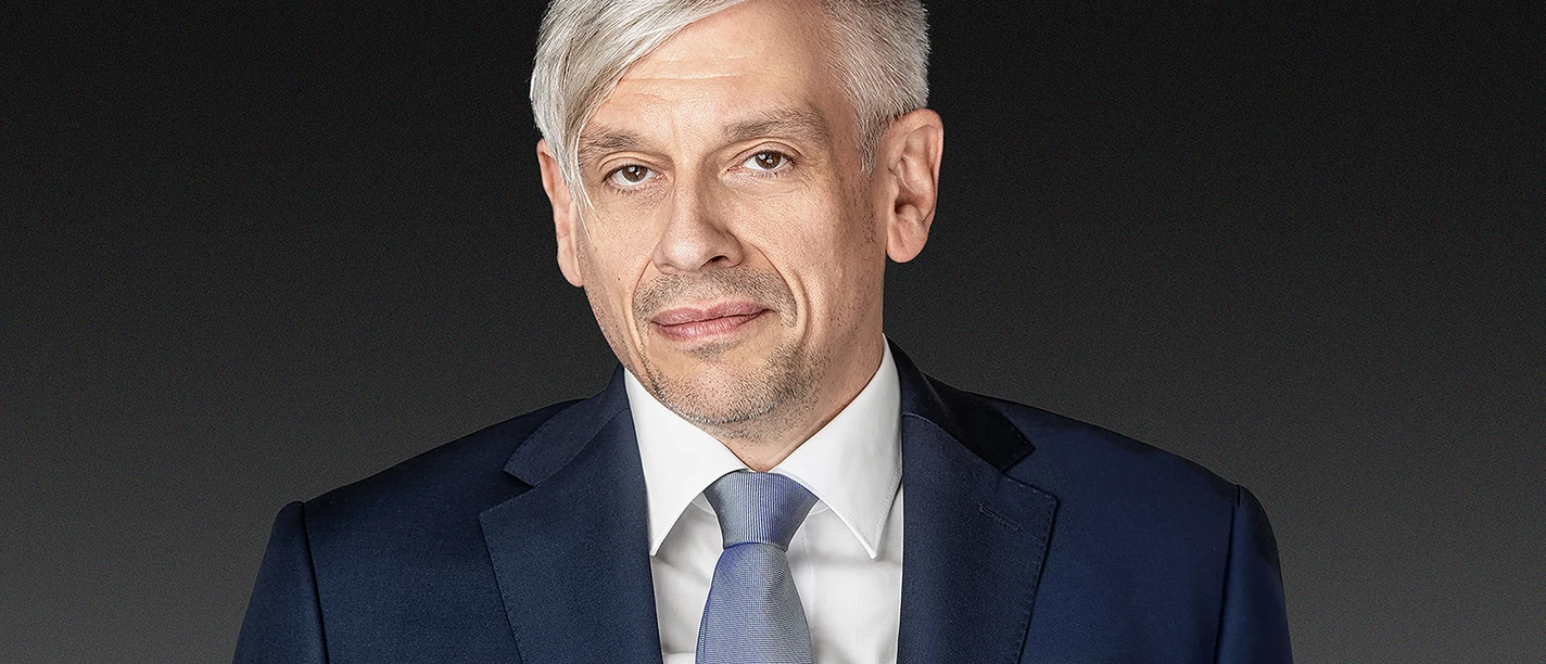 Mathias Tretter Souveraen Porträt eines Mannes mit grauem Haar im dunklen Anzug und hellblauer Krawatte vor dunklem Hintergrund.