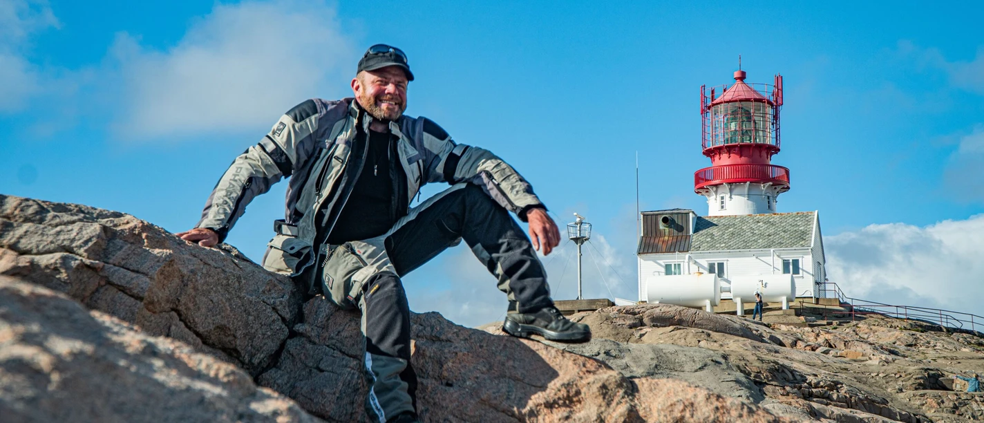Mann in Outdoor-Kleidung sitzt auf Felsen vor einem Leuchtturm an einer felsigen Küste.