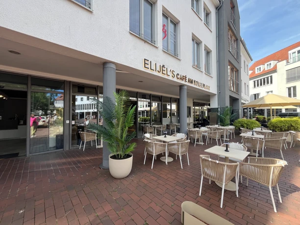 Außenbereich von Elijel's Café am Kolbeplatz mit weißen Tischen und Palmen auf der Terrasse.