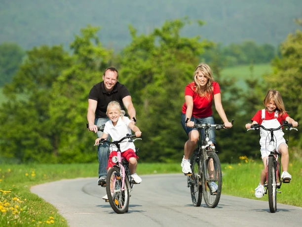 Radfahrende Familie Familie mit zwei Kindern radelt lächelnd auf einem ländlichen Weg entlang grüner Wiesen.