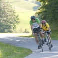 Rasante Abfahrt Zwei Radfahrer in sportlicher Haltung fahren eine kurvige Landstraße hinunter, umgeben von Bäumen.