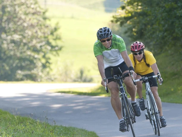 Rasante Abfahrt Zwei Radfahrer in sportlicher Haltung fahren eine kurvige Landstraße hinunter, umgeben von Bäumen.