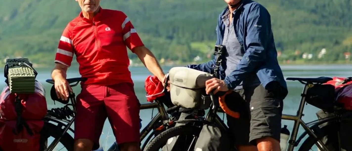 Zwei Radreisende mit bepackten Fahrrädern vor See und grünen Bergen.
