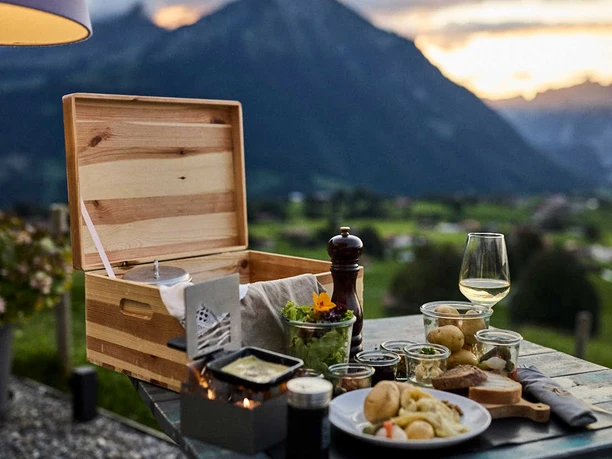 A unique wooden box containing a tabletop raclette set, potatoes, bread, wine, and Swiss specialties, served on the terrace with a panoramic view of the Niesen