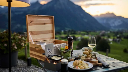 Caisse en bois originale contenant un service de raclette, des pommes de terre, du pain, du vin et des spécialités suisses, à déguster sur la terrasse face au panorama du Niesen