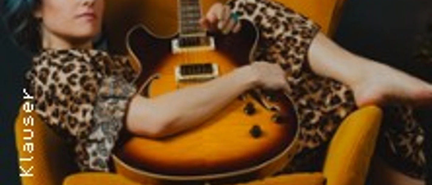 Charly Klauser mit Gitarre auf orangefarbenem Sessel vor dunklem Hintergrund