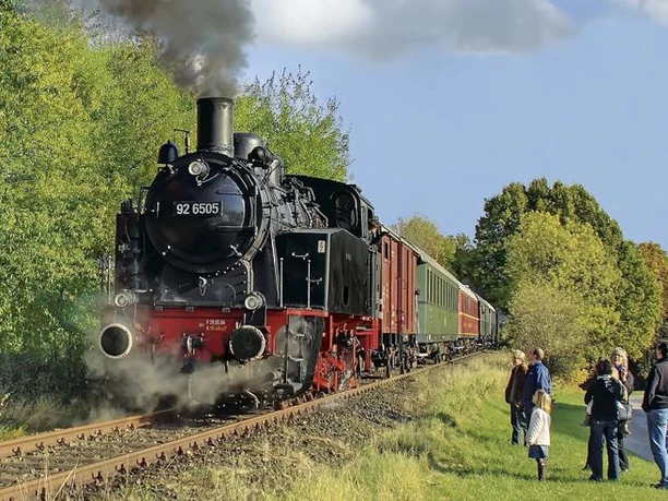 Museumseisenbahn Eine Dampflokomotive fährt durch eine grüne Landschaft, mehrere Menschen beobachten sie am Gleisrand.