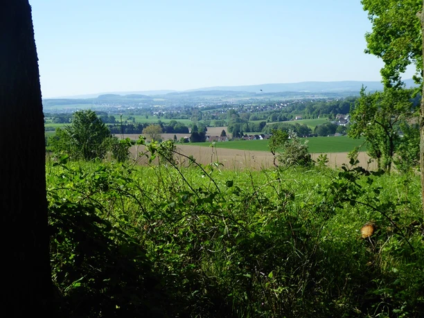 Blick über Felder und Wiesen auf eine weite Landschaft mit Hügeln und einem Dorf im Hintergrund.
