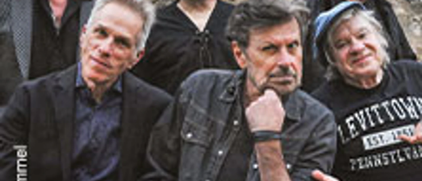 Sechs Musiker der Band The Hooters vor Steinmauer mit weißem Logo