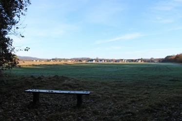 Bank am Bärental; Blickrichtung Ost Holzbank auf einer Wiese vor einer weiten Landschaft; Blick auf Gebäude am Horizont unter blauem Himmel.