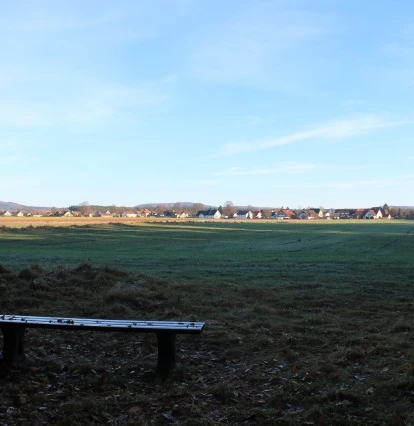 Bank am Bärental; Blickrichtung Ost Holzbank auf einer Wiese vor einer weiten Landschaft; Blick auf Gebäude am Horizont unter blauem Himmel.