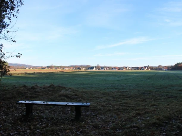 Bank am Bärental; Blickrichtung Ost Holzbank auf einer Wiese vor einer weiten Landschaft; Blick auf Gebäude am Horizont unter blauem Himmel.