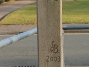 Wanderzeichenpfahl Holzpfahl mit Eulen-Gravur und Richtungsanzeige für Wanderer vor einem Wohnhaus an einer Straße.