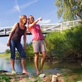 Zwei Frauen stehen barfuß in einem flachen Fluss und schauen lächelnd in die Ferne, im Hintergrund eine Brücke.