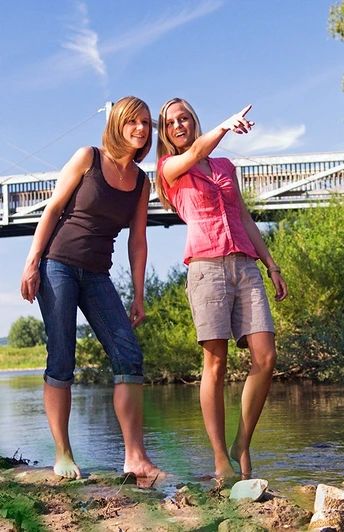Zwei Frauen stehen barfuß in einem flachen Fluss und schauen lächelnd in die Ferne, im Hintergrund eine Brücke.