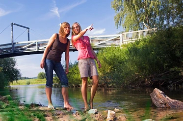 Nethemündung Zwei Frauen stehen barfuß in einem flachen Fluss und schauen lächelnd in die Ferne, im Hintergrund eine Brücke.