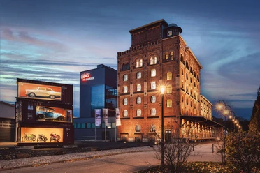 Außenansicht PS.SPEICEHR Beleuchtetes PS.SPEICHER-Gebäude bei Dämmerung mit Oldtimern in Glascontainern.