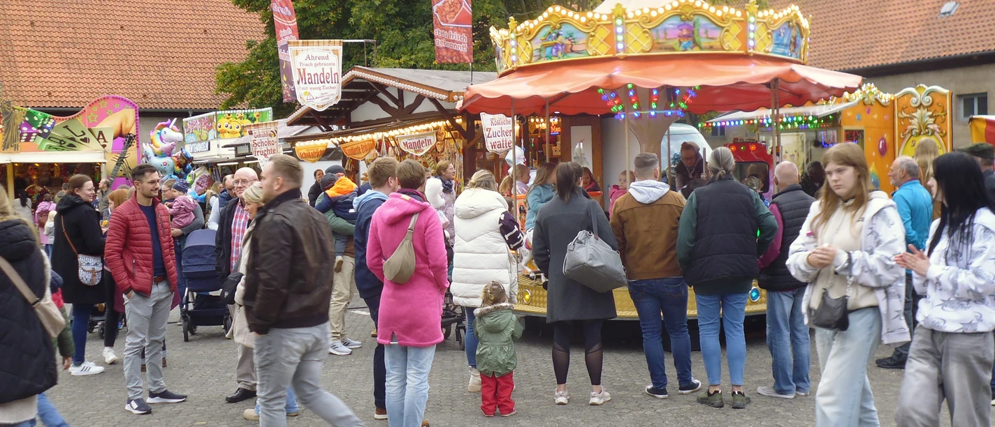 Menschen auf einem Jahrmarkt mit buntem Kinderkarussell und Mandelbude im Hintergrund.