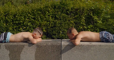 Two boys are lying face down at the edge of the pool, looking at each other across a green hedge