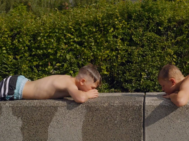 Two boys are lying face down at the edge of the pool, looking at each other across a green hedge