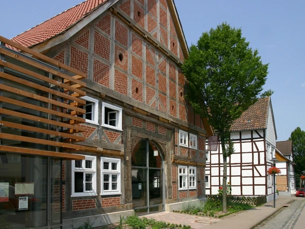 Ein traditionelles Fachwerkhaus mit Backsteinmusterung steht neben modernem Glasbau unter blauem Himmel.