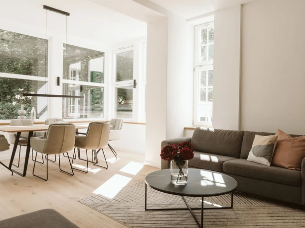 Villa am Stadtpark Heller Wohn- und Essbereich mit grauem Sofa, Rundtisch und Esstisch vor großen Fenstern.