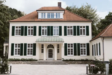 Weiße Villa am Stadtpark mit grünen Fensterläden und rotem Ziegeldach.