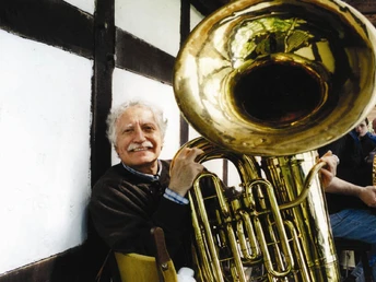 Ein älterer Mann spielt fröhlich eine goldene Tuba in einem Fachwerkgebäude.