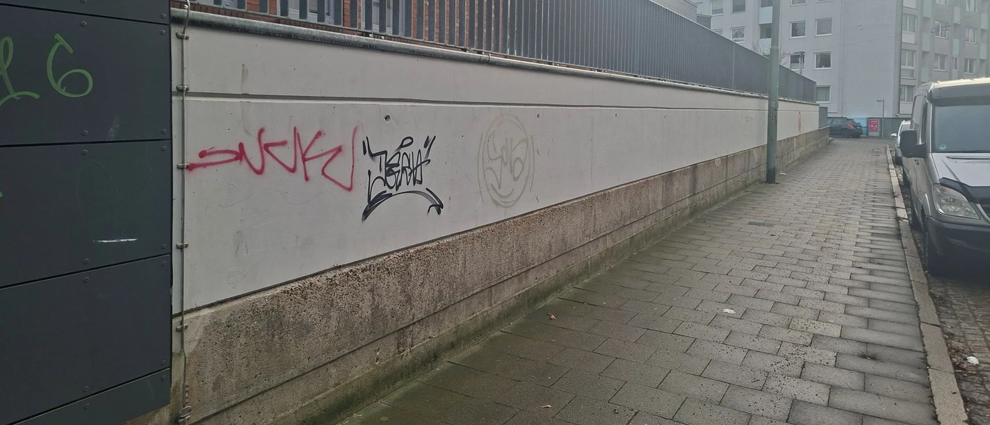 Bild2 Leere Wand (c) Lehe-Treff_WEB.jpg Graffiti an langer Betonwand neben Gehweg und Backsteingebäude bei grauem Himmel.