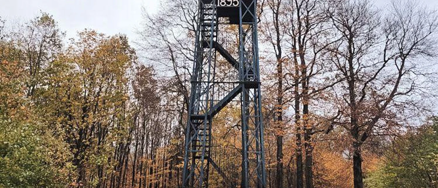 Ein Aussichtsturm aus dem Jahr 1895 steht in einem herbstlichen Wald. Die Umgebung ist mit buntem Laub geschmückt.
